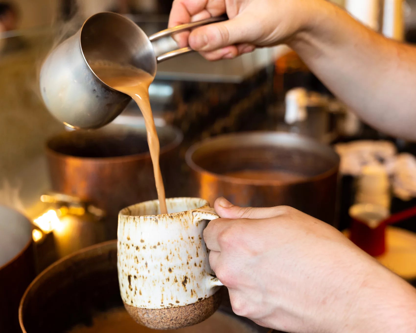 A hand pouring hot tea from a carafe into a ceramic mug, with a cozy kitchen background.
