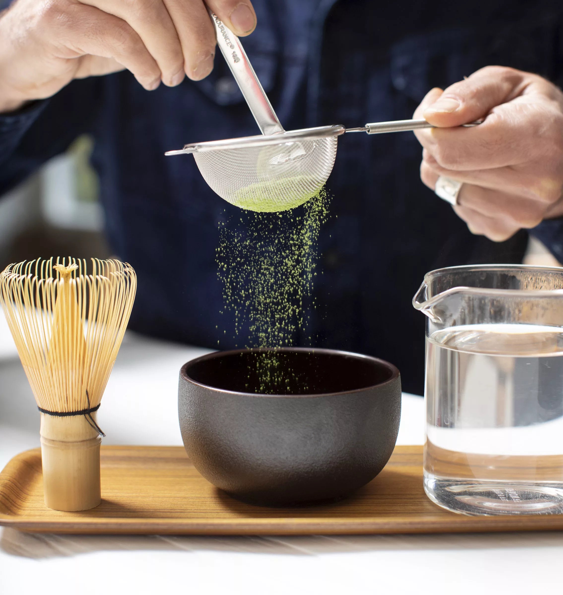 A person pours green tea from a teapot into a black matcha bowl.