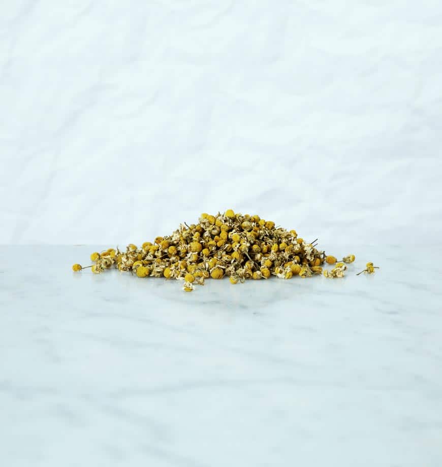 A pile of dried chamomile flowers on a white surface, ready for use in herbal tea preparation.